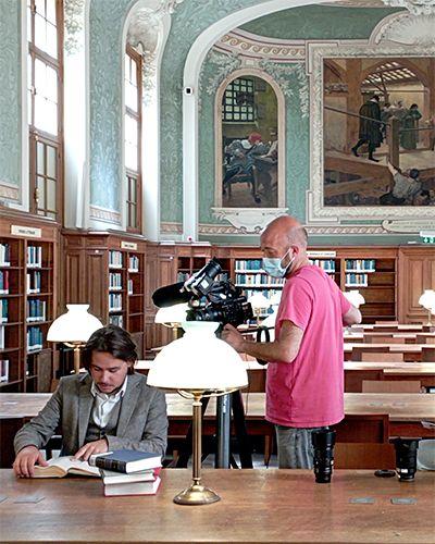 Équipe de tournage de trois personnes, un homme assis à une table lisant, un autre le filmant avec une caméra sur pied, une femme appuyée contre la table regarde la scène