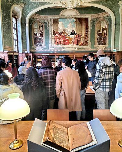 Un groupe de lycéens debout dans la salle Romilly, un manuscrit médiéval est présenté ouvert sur une table au premier plan