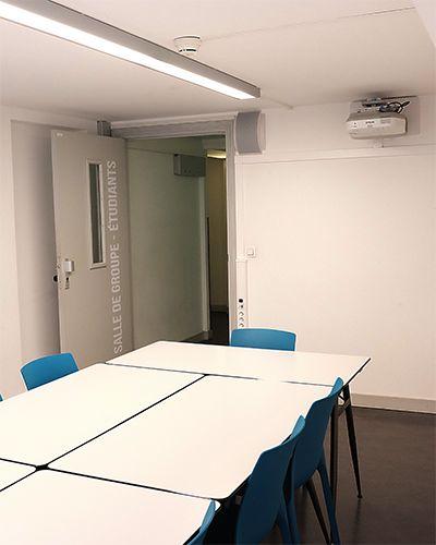 Salle de travail avec une grande table blanche, des chaises bleues et un vidéoprojecteur