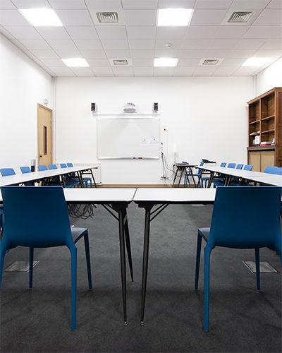 Salle avec des tables blanches installées en U, des chaises bleues et un tableau blanc avec vidéoprojecteur